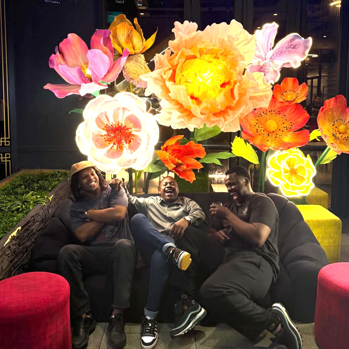 Three friends laughing on a lounge sofa beneath giant illuminated blooms