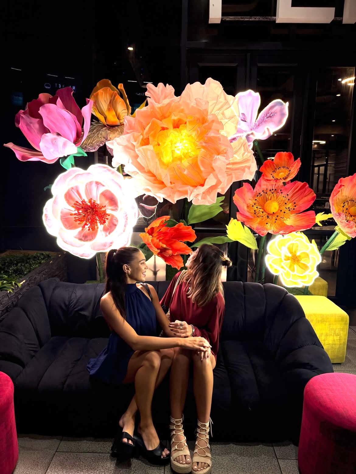 Two women seated beneath a wall of giant glowing flowers at night